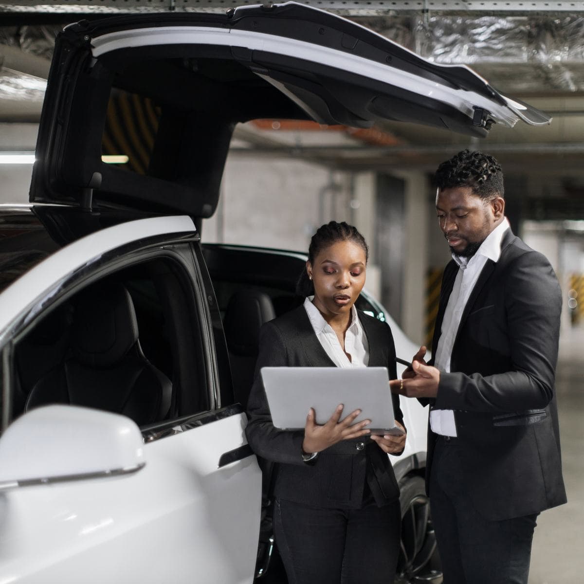 Two fleet professionals reviewing information on a laptop beside a white electric vehicle in a parking garage. Two fleet professionals reviewing information on a laptop beside a white electric vehicle in a parking garage.