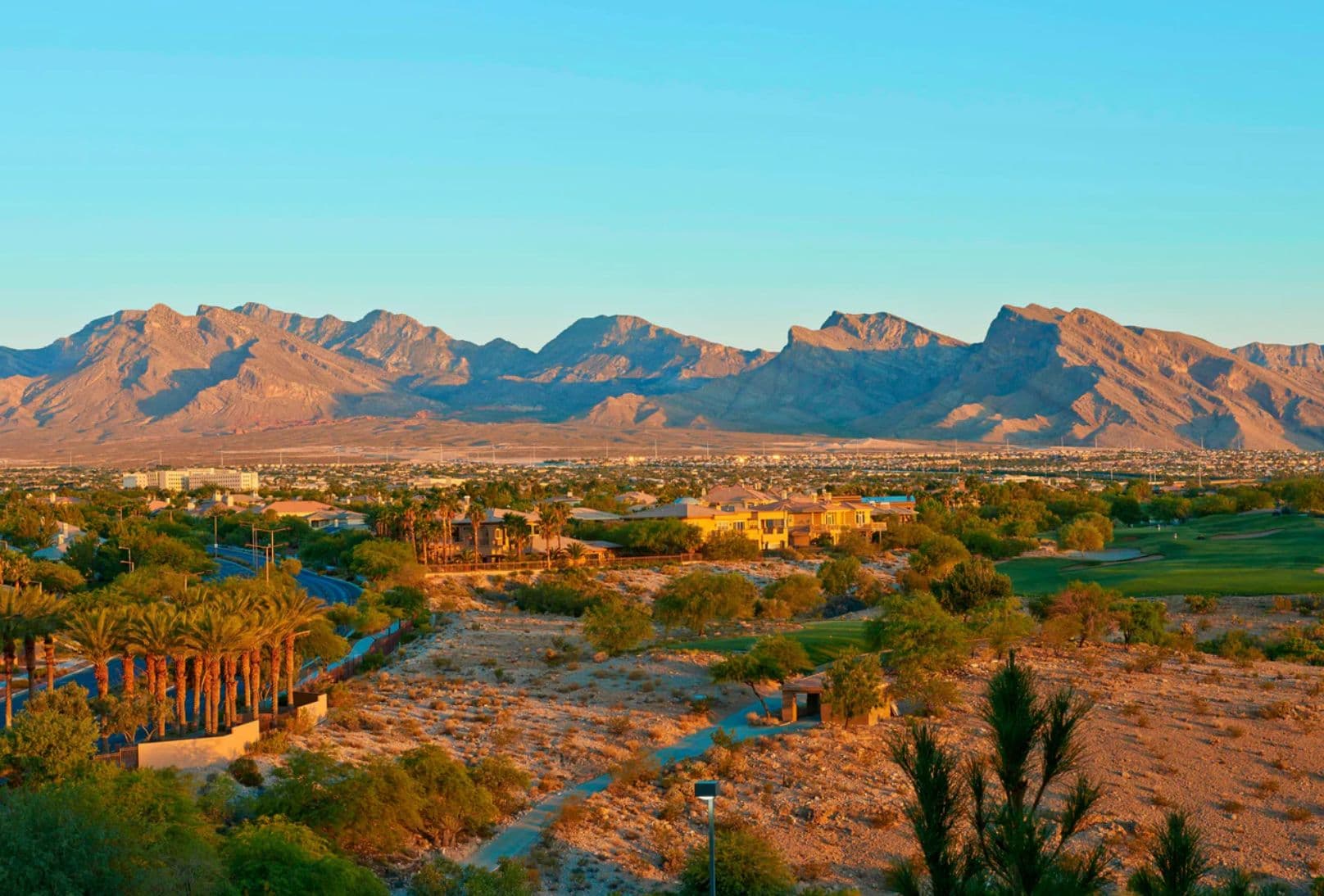 Scenic landscape view of the Las Vegas valley at sunset, with desert mountains, palm trees, homes, and green golf course terrain in the foreground. Scenic landscape view of the Las Vegas valley at sunset, with desert mountains, palm trees, homes, and green golf course terrain in the foreground.