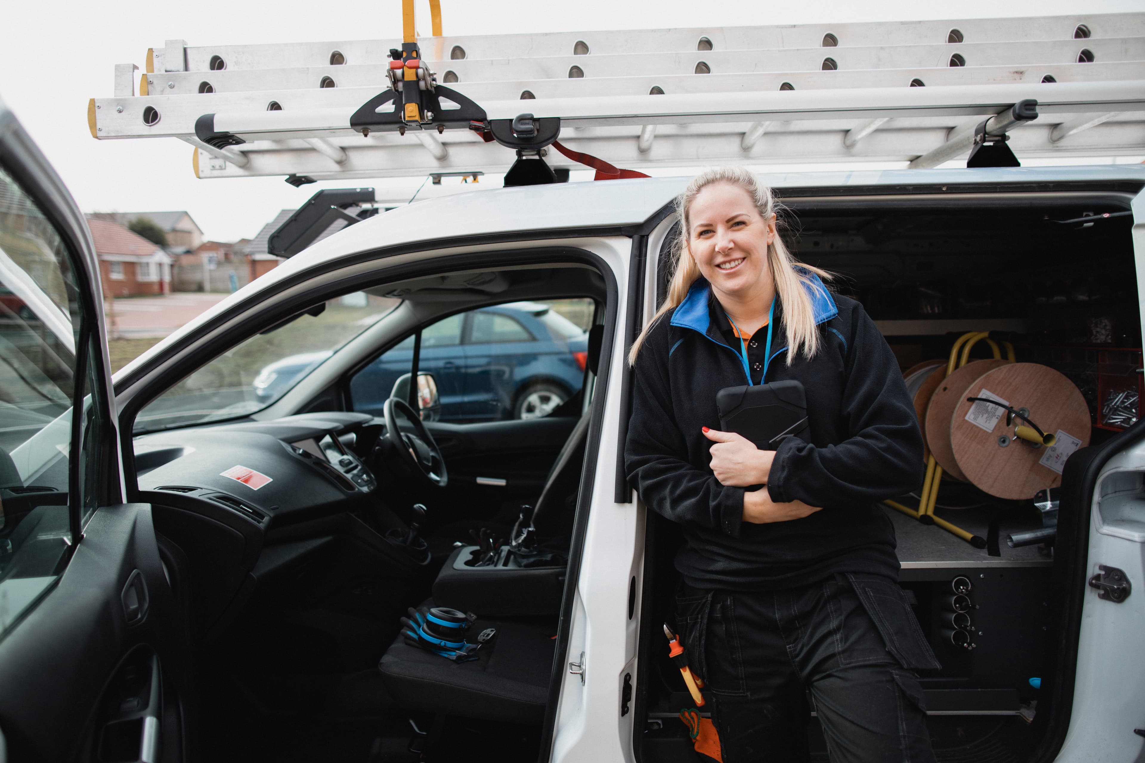 Vehicle upfitting shown in a fully equipped work van with organized shelving, cable reels, and a roof ladder rack, beside a service technician. Vehicle upfitting shown in a fully equipped work van with organized shelving, cable reels, and a roof ladder rack, beside a service technician.