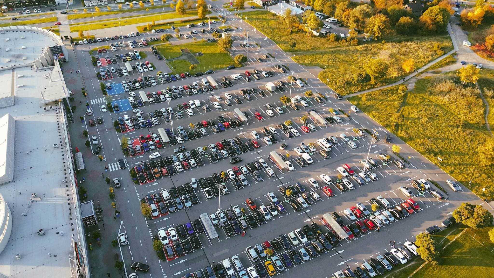 Aerial view of a large parking lot filled with parked vehicles beside a commercial building, surrounded by roads and autumn trees. Aerial view of a large parking lot filled with parked vehicles beside a commercial building, surrounded by roads and autumn trees.
