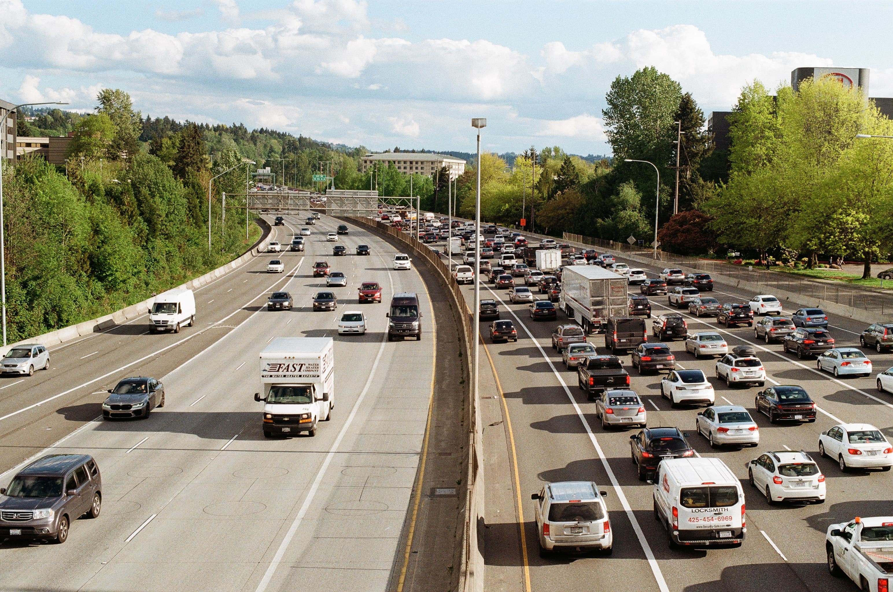 A divided highway with heavy traffic on one side and lighter traffic on the other, surrounded by green trees under a partly cloudy sky. A divided highway with heavy traffic on one side and lighter traffic on the other, surrounded by green trees under a partly cloudy sky.