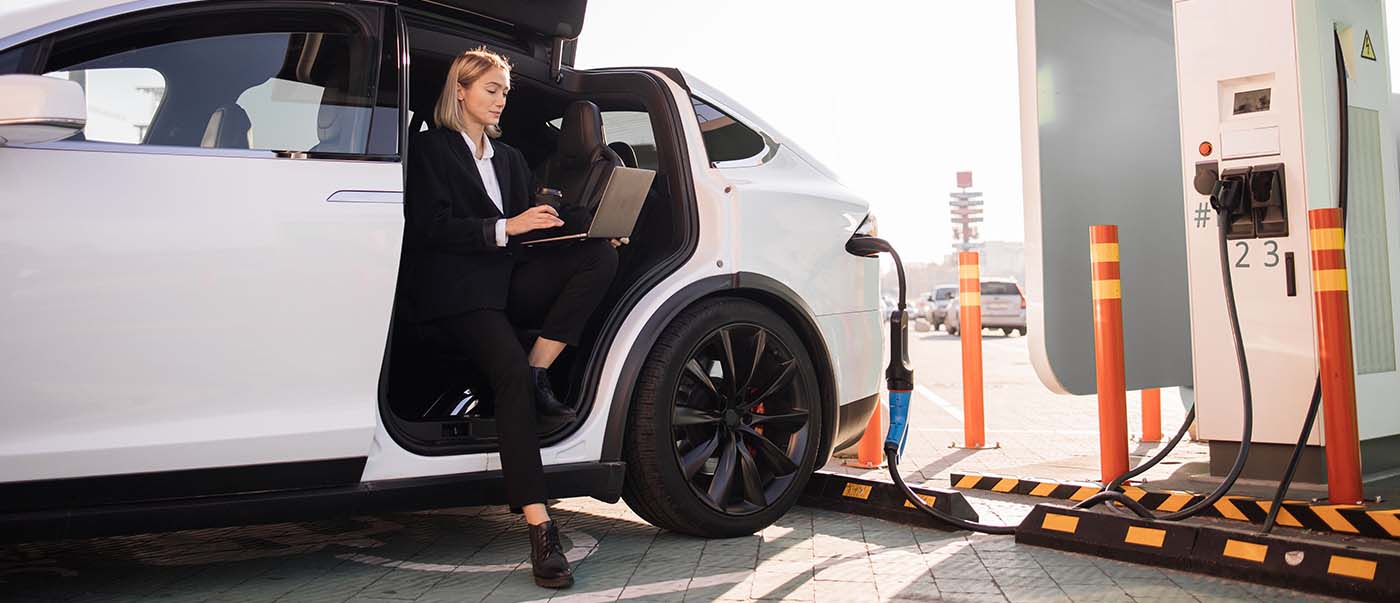 Businesswoman using a laptop while charging an electric vehicle at a public EV charging station. Businesswoman using a laptop while charging an electric vehicle at a public EV charging station.