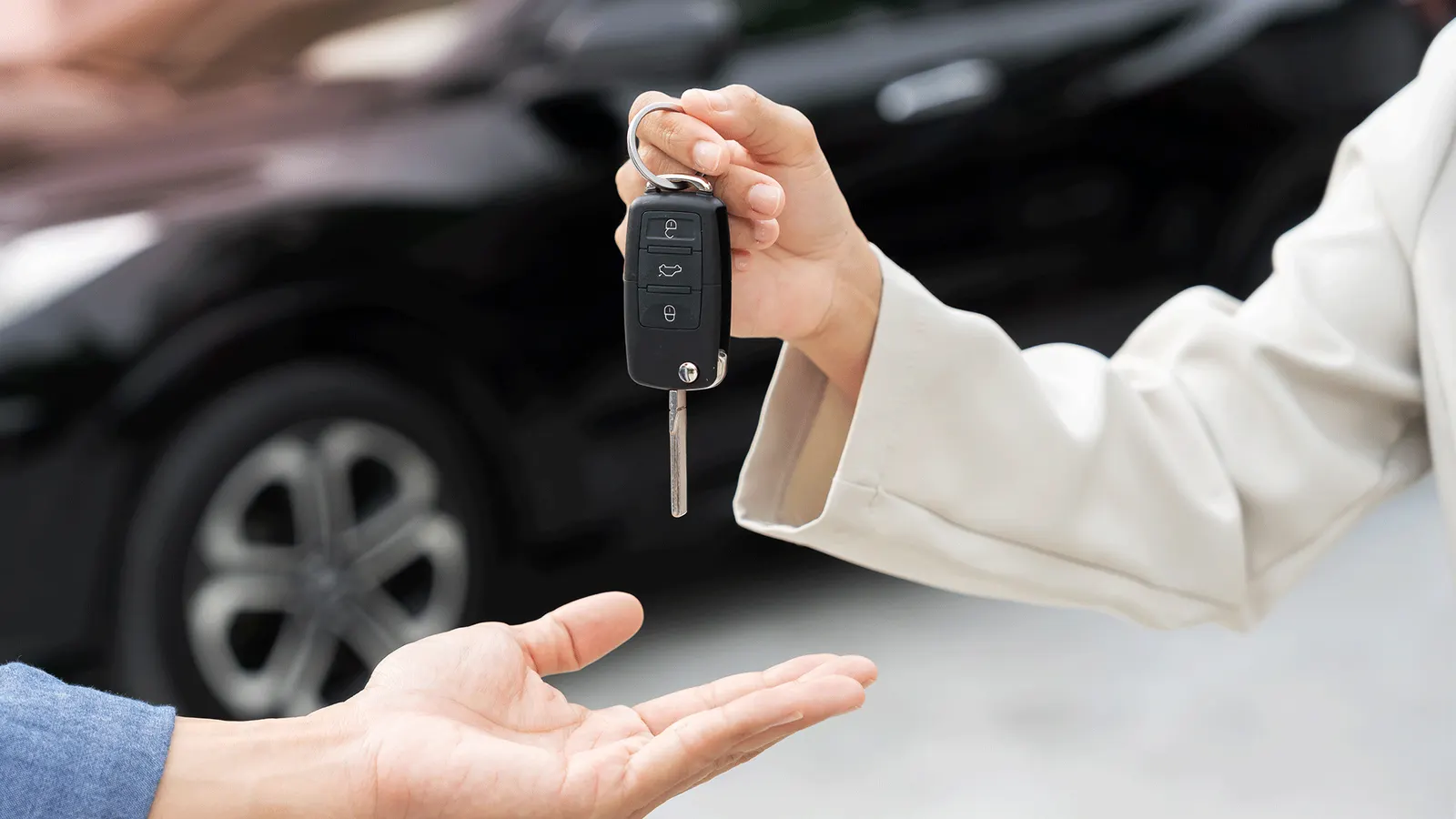 Close-up of a person handing over car keys to another person in front of a black vehicle. Close-up of a person handing over car keys to another person in front of a black vehicle.
