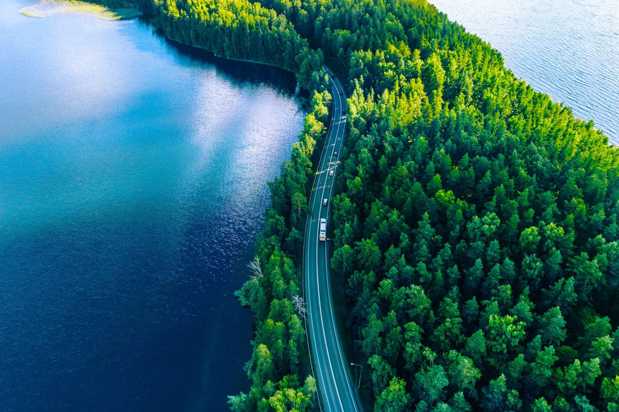 Aerial view of a winding road through a lush green forest beside a blue lake. Aerial view of a winding road through a lush green forest beside a blue lake.