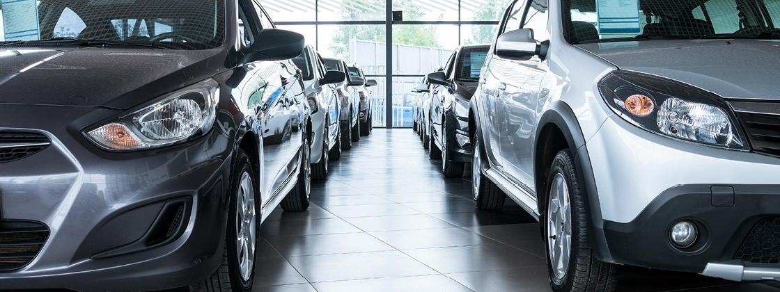 A row of cars parked inside a showroom with large windows in the background, allowing natural light to illuminate the space. The cars are lined up closely together, showcasing various models and colors.