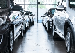 A row of cars parked inside a showroom with large windows in the background, allowing natural light to illuminate the space. The cars are lined up closely together, showcasing various models and colors.