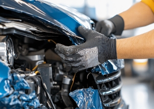 A mechanic works on the damaged hood of a blue sedan that has been in a fleet collision.